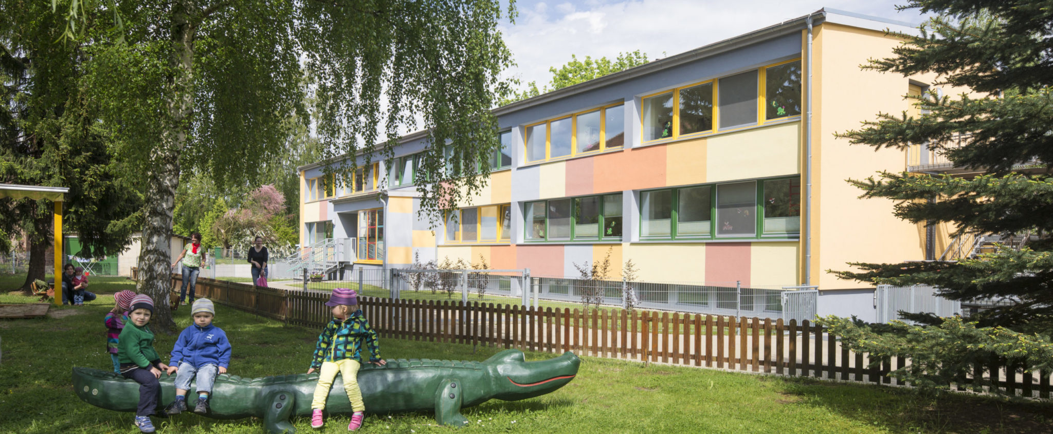 Blick auf den Garten und das Gebäude, im Vordergrund sitzen drei Kinder auf einem Holzkrokodil