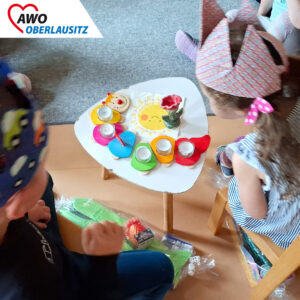 zwei Kinder mit Kronen sitzen vor einem Tisch. Auf diesem sind Teelichter platziert.