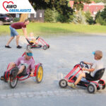 Zwei Kinder fahren mit Tretautos. Ein kleineres Kind wird von einem Erwachsenen Angeschoben