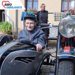 Nahaufnahme von einem Mann der in dem Beiwagen eines Motorrades sitzt. Im Hintergrund steht ein weiterer Mann