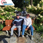 Mann und Frau sitzen auf einer Bank. Im Hintergrund sind Pflanzen und ein Motorrad