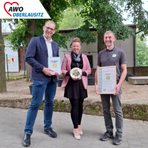 Zwei Männer und eine Frau in der Mitte lachen in die Kamera. Sie halten die Ernennungsurkunde zur Naturpark-Kita in den Händen und die Plakette für die Kita.
