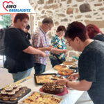 Großer Tisch mit vielfältiger Kuchenauswahl. Besucher kaufen Kuchen.
