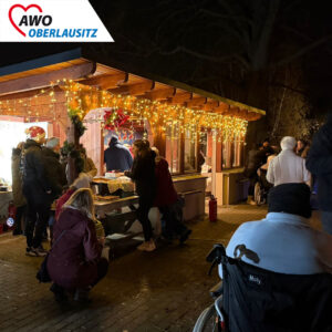 Weihnachtliche Holzbuden mit Lichterketten geschmückt, Senioren im Rollstuhl, warm eingepackt mit Pflegekräften, Besucher, Angehörige und Anwohner sind zu sehen. Sie genießen die weihnachtliche Stimmung.