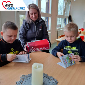 Eine Mitarbeiterin der Firma Konzmann sitzt mit eienr Jacke d er Firma Konzmann und einer Kiste, auf welcher Konzmann steht am Tisch. Neben ihr zwei Vorschuljungs mit einem Pixibuch in der Hand.