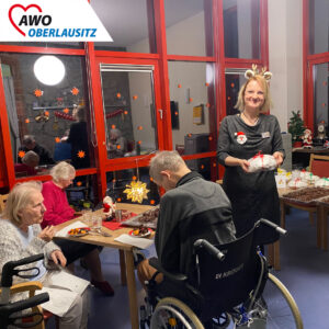 Senioren sitzen beim Adventskaffee mit Leckereien vor sich auf dem Tisch. Eine Mitarbeiterin mit Elchgeweih und im Glitzerkleid steht im Raum und strahlt in die Kamera.