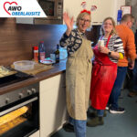 Zwei Mitarbeiterinnen mit lustigem Weihnachtshaarreif und roter Schürze stehen in der Küche des Altenheims beim Plätzchen backen und winken fröhlich.