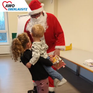 Ein etwa 8- bis 9-jähriges Mädchen hat einen kleinen Jugen auf den Arm. Beide stehen vpr dem Weihnachtsmann.