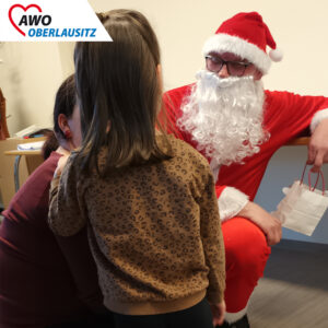 Ein kleines Mädchen hält sich an der neben ihm hockenden Mutter fest. Vor ihnen hockt ebensfalls der Weihnachtsmann mit einer kleinen Geschenktüte in der Hand.