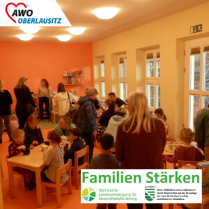 In einem hellen freundlichen Raum sind viele Eltern und Kinder versammelt. Es wird geredet an Tischen gemeinsam gespielt.
