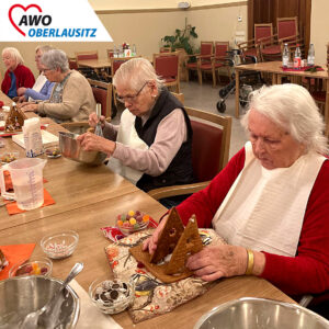 Fünf Seniorinnen sitzen an langen Tischen in einem großen Saal. Eine Frau rührt in einer Edelstahlschüssel den Zuckerguss, eine andere klebt die Wände eines Lebkuchenhauses zusammen. Die hinteren Damen dekorieren fleißig und sitzen beinander. Überall auf den Tischen stehen kleine Schüsselchen mit Streuseln, Gummibären und Schokotalern.