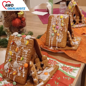 Auf einem weihnachtlich dekorierten Tisch stehen drei fertige Lebkuchenhäuser. Auf den Dächern kleben dank Zuckerguss große bunte Smarties, Gummibärchen und Schokotaler. Vor den Häuschen stehen stehen jeweils drei kleine Lebkuchenmännlein.