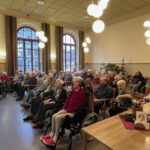 In einem großen weihnachtlich geschmückten Saal mit vielen hohen Fenstern sitzen viele Senioren, Männer und Frauen, auf Stühlen oder in Rollstühlen und lauschen aufmerksam dem Chor.