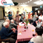 In einem großen geschmückten Gemeinschaftsraum oder -saal feiern ausgelassen Senioren und Seniorinnen sowie Mitarbeiter einen zünftigen Baudenabend. Die Senioren an den Tischen klatschen zur Musik. Hinter den Tischen laufen Bewohner und Mitarbeit in einer Art Polonaise. Auf Tischen mit rot-weiß-karierten Tischdecken stehen Getränke und frische Blumen sowie Dekoration. Alle haben fröhliche. lachende Gesichter.