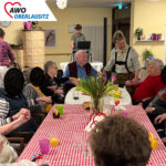 In einem großen geschmückten Gemeinschaftsraum oder -saal feiern ausgelassen Senioren und Seniorinnen sowie Mitarbeiter einen zünftigen Baudenabend. Die Senioren an den Tischen klatschen zur Musik. Hinter den Senioren ist ein DJ zu sehen. Auf Tischen mit rot-weiß-karierten Tischdecken stehen Getränke und frische Blumen sowie Dekoration. Alle haben fröhliche. lachende Gesichter.