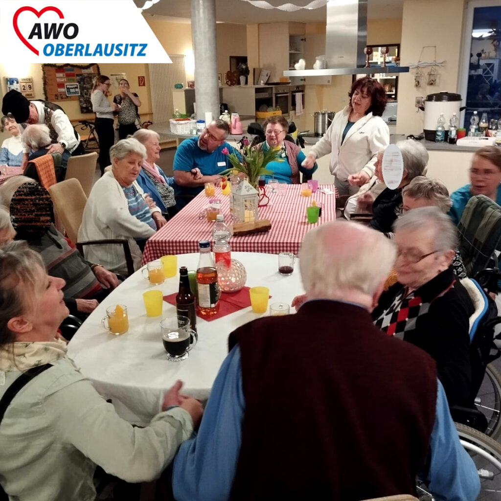 In einem großen geschmückten Gemeinschaftsraum oder -saal feiern ausgelassen Senioren und Seniorinnen sowie Mitarbeiter einen zünftigen Baudenabend. Die Senioren an den Tischen klatschen zur Musik. Hinter den Tischen laufen Bewohner und Mitarbeit in einer Art Polonaise. Auf Tischen mit rot-weiß-karierten Tischdecken stehen Getränke und frische Blumen sowie Dekoration. Alle haben fröhliche. lachende Gesichter.
