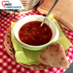 Auf der rot-weiß-karierten Tischdecke steht eine appetitlich aussehende Gulaschsuppe in einer weißen Schüssel. Daneben liegt frisches Brot.