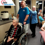 EIn fröhlich mitsingender Mitarbeiter schiebt eine Bewohnerin im Rollstuhl während der Polonaise durch den Saal. An seinen Schultern hinter ihm geht lächelnd eine weitere Bewohnerin.