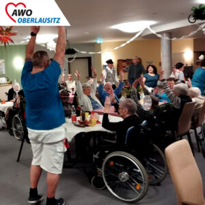 In einem großen geschmückten Gemeinschaftsraum oder -saal feiern ausgelassen Senioren und Seniorinnen sowie Mitarbeiter einen zünftigen Baudenabend. Die Hände sind stimmungsvoll zur Musik nach oben gerichtet. Auf Tischen mit rot-weiß-karierten Tischdecken stehen Getränke. Alle haben fröhliche. lachende Gesichter.