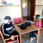 Zwei Schülerinnen sitzen an einem Tisch. Sie haben je eine VR-Brille auf und sehen sich im Raum um.