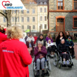 Im Außenbereich auf dem Hof des Altenpflegeheims stehen die Schülerinnen jeweils hinter einem Rollstuhl mit warm abgezogenen Bewohnern. Im Vordergrund sind noch zwei Mitarbeiterinnen mit roten "AWO Oberlausitz"-Jacken zu sehen.