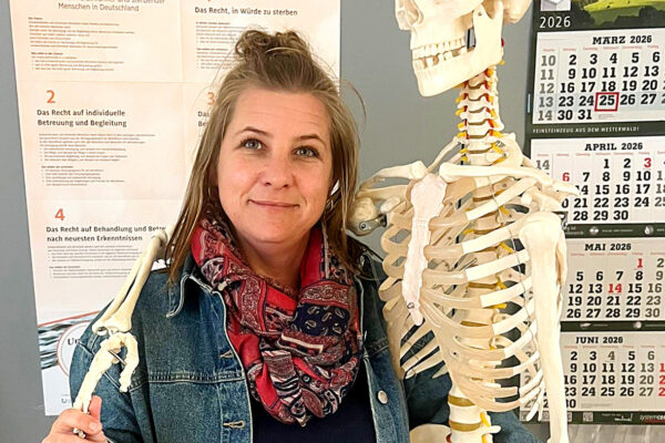 Eine Frau mit halblangen braunen Haaren und in Jeansjacke steht mit einem Skelett, welches den Arm um sie gelegt hat in einem Ausbildungskabinett der theoretischen Berufsausbildung.