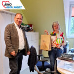 Geschäftsführer Dirk Reinke steht breit lächelnd im Verwaltungsbüro des Altenpflegeheimes und hat die Jubiläumstüte gerade an die dort arbeitende Kollegin übergeben. Freudig lächelnd hält diese die Tüte in der Hand.