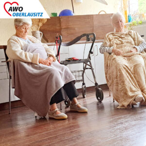 Zwei Seniorinnen sitzen bequem mit Sohlfühldecke in einem Stuhl. Die Hände im Schoß verschränkt und die Augen geschlossen.